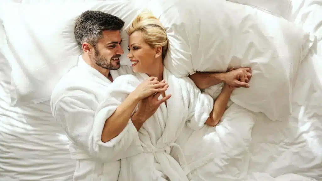 A smiling man and woman wearing white bathrobes are lying down on a white bed.