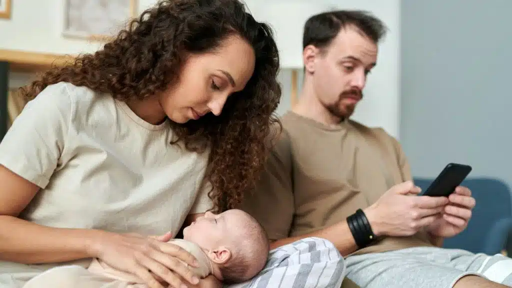 A woman lovingly looks down at the baby in her lap while a man next to her looks at his smartphone.