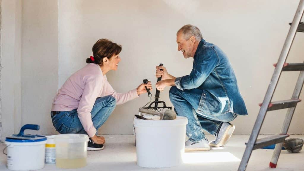 Two smiling people kneeling and working together with a paint roller and bucket.
