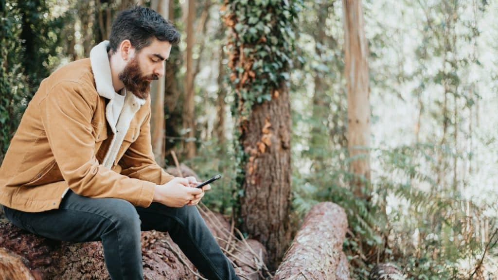 A bearded man using his phone outdoors while sitting on a log.