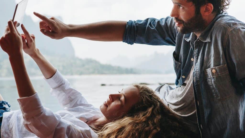 A couple relaxes outdoors near water, with the woman lying down and holding a tablet while the man points towards it.