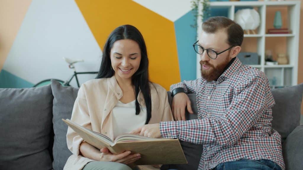 A smiling couple sits on a couch, looking at and pointing into a photo album.