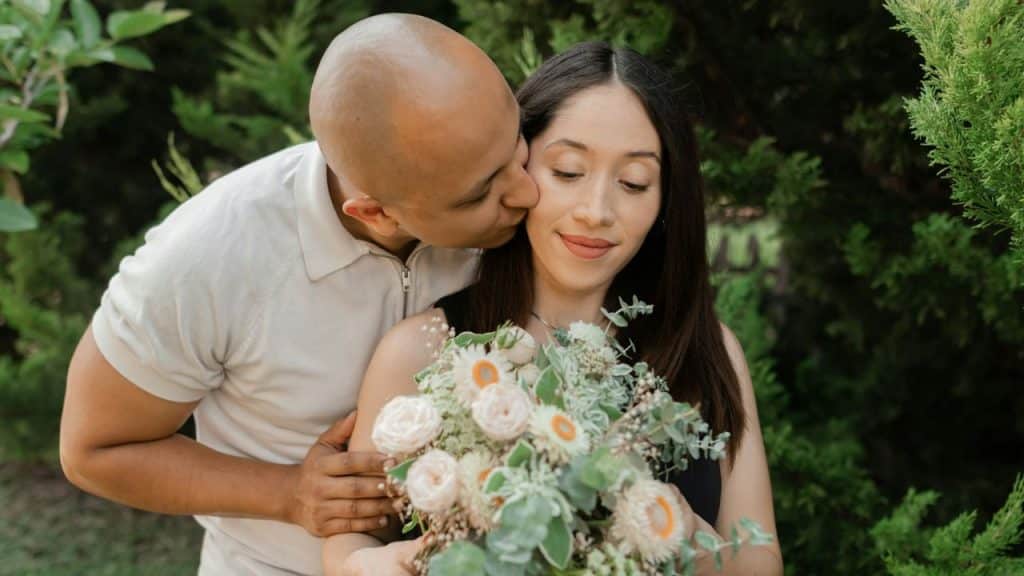 A bald man in a beige shirt kisses a woman's cheek while she holds a bouquet of flowers.