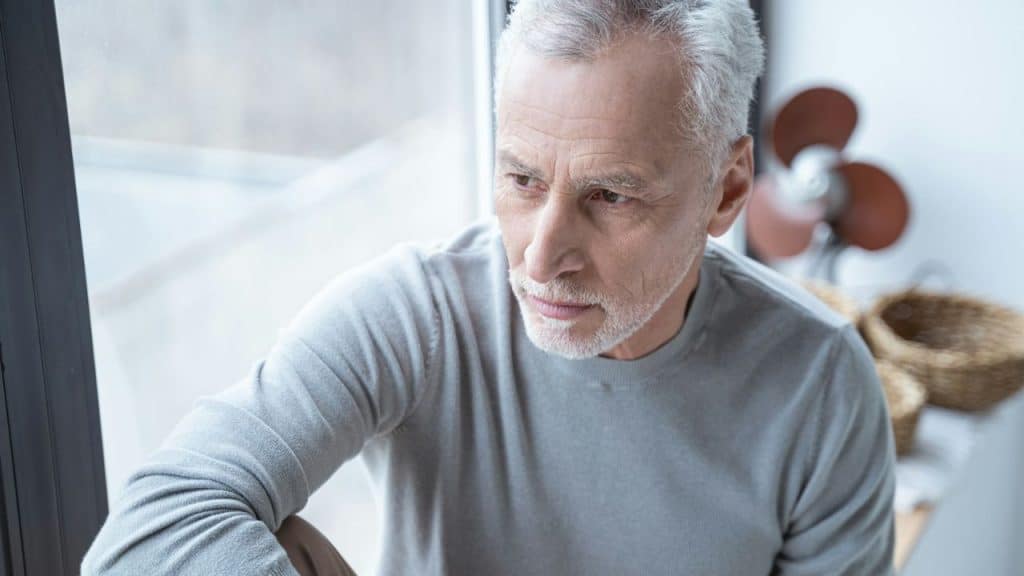 A thoughtful, older man with gray hair and a beard looks out a window.
