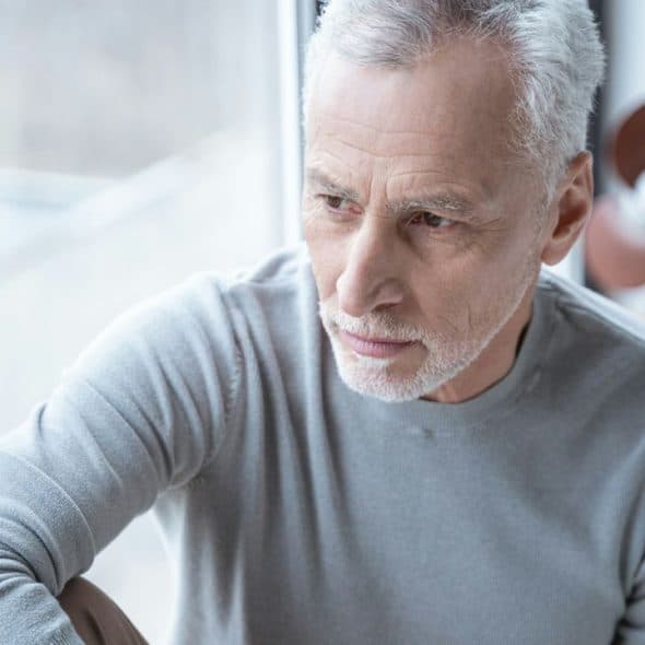 A thoughtful, older man with gray hair and a beard looks out a window.