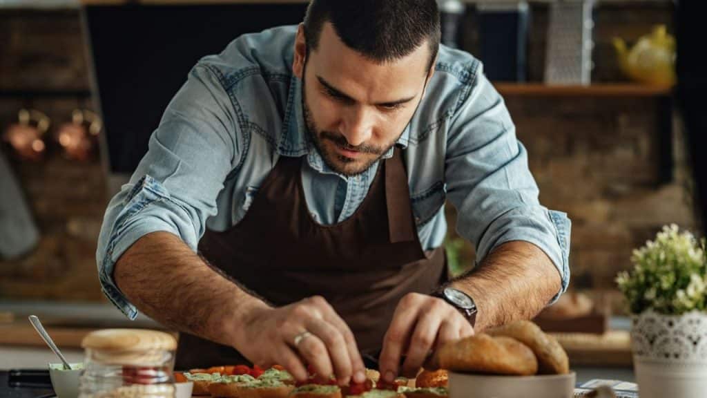 A focused man in a denim shirt and apron is preparing food on a kitchen counter.