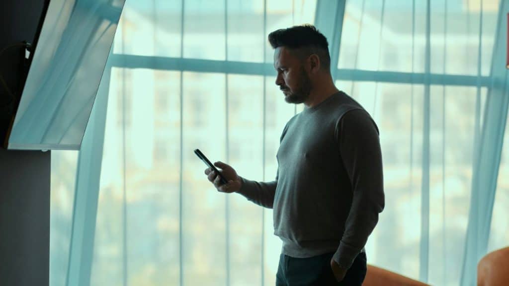 A pensive man is looking at his phone in front of a bright, sheer curtained window.
