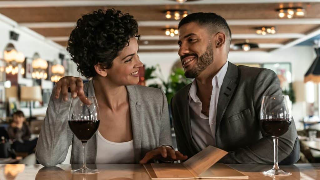 Smiling couple at a bar or restaurant, looking at each other over wine glasses and a menu.