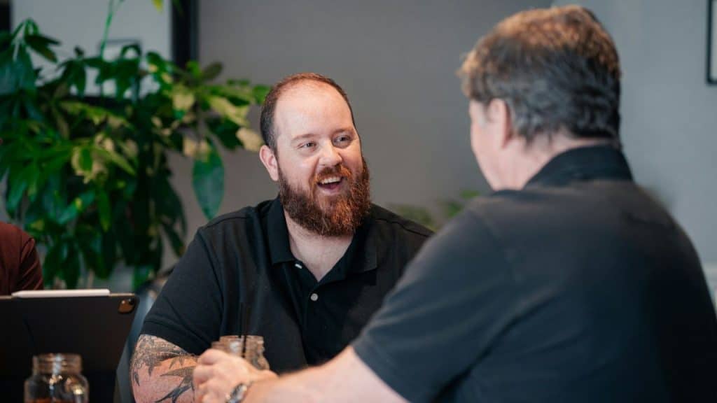 Smiling man with a bald head and a full beard talking to another man across a table.