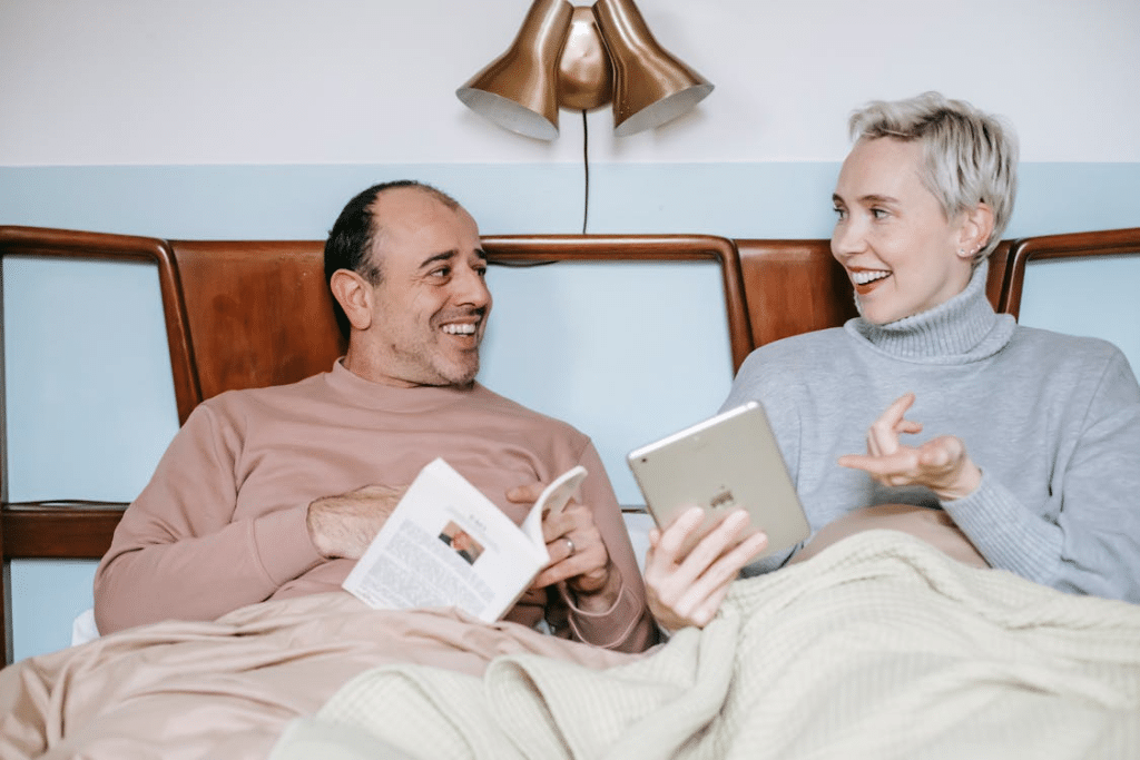 Cheerful diverse couple relaxing in bed with book and tablet during lazy weekend