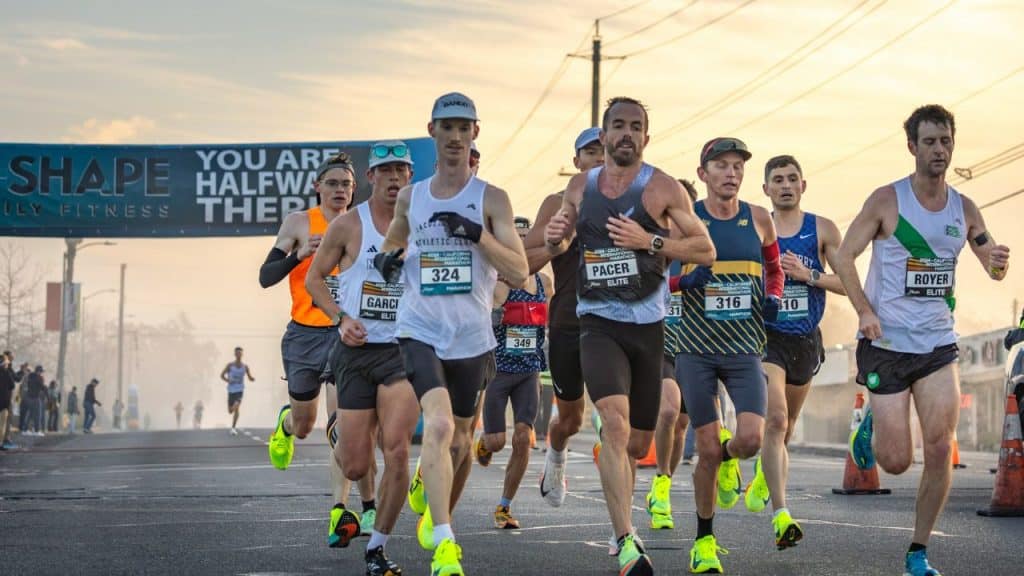 A group of male runners with bibs is finishing a race under a banner.