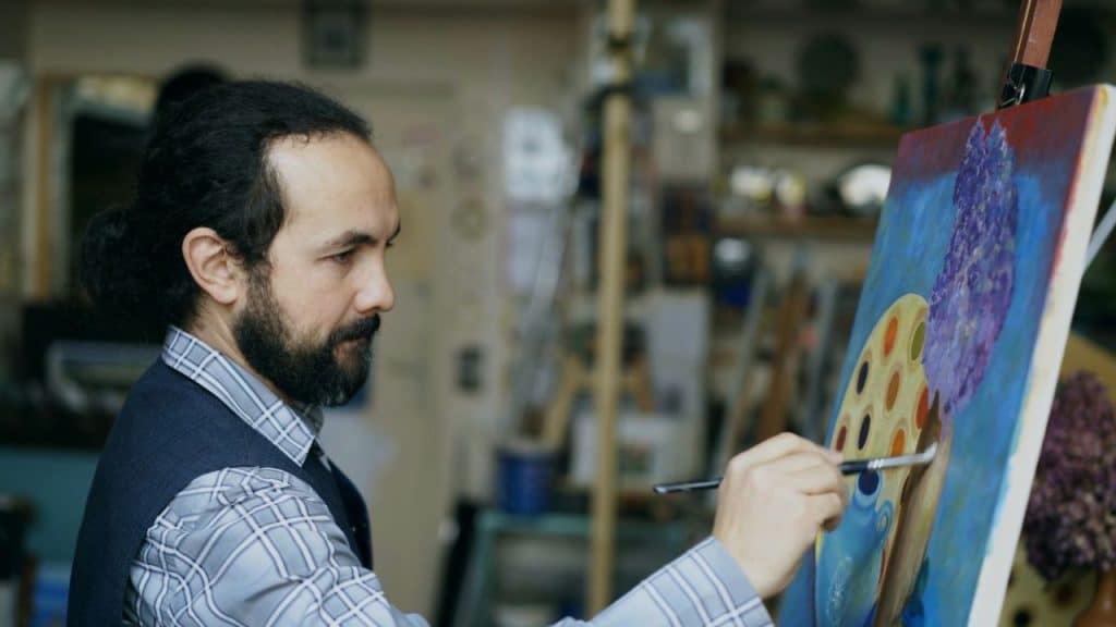 A focused, bearded male artist in a vest and plaid shirt paints on a canvas in his studio.