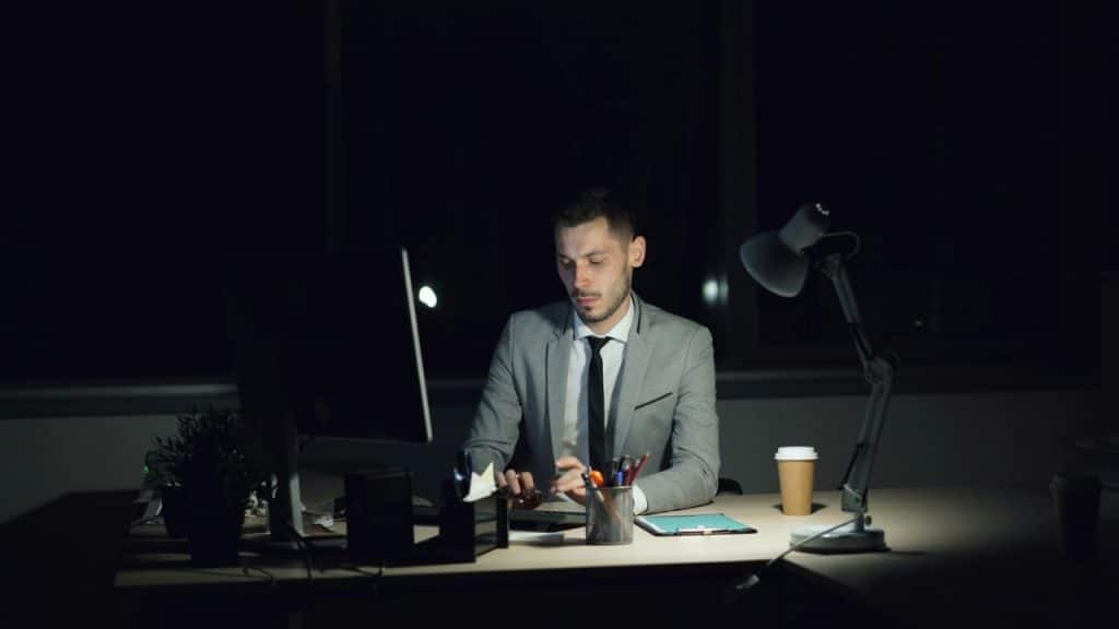 A man in a gray suit is working at a desk in a dark office at night.