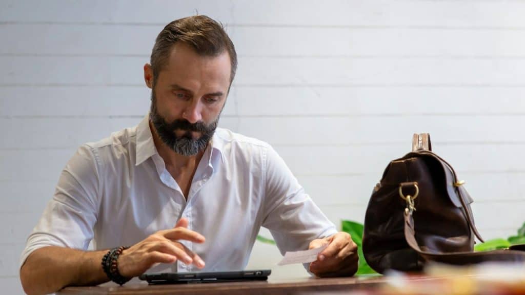 Bearded man in white shirt looking at a small card and a tablet.
