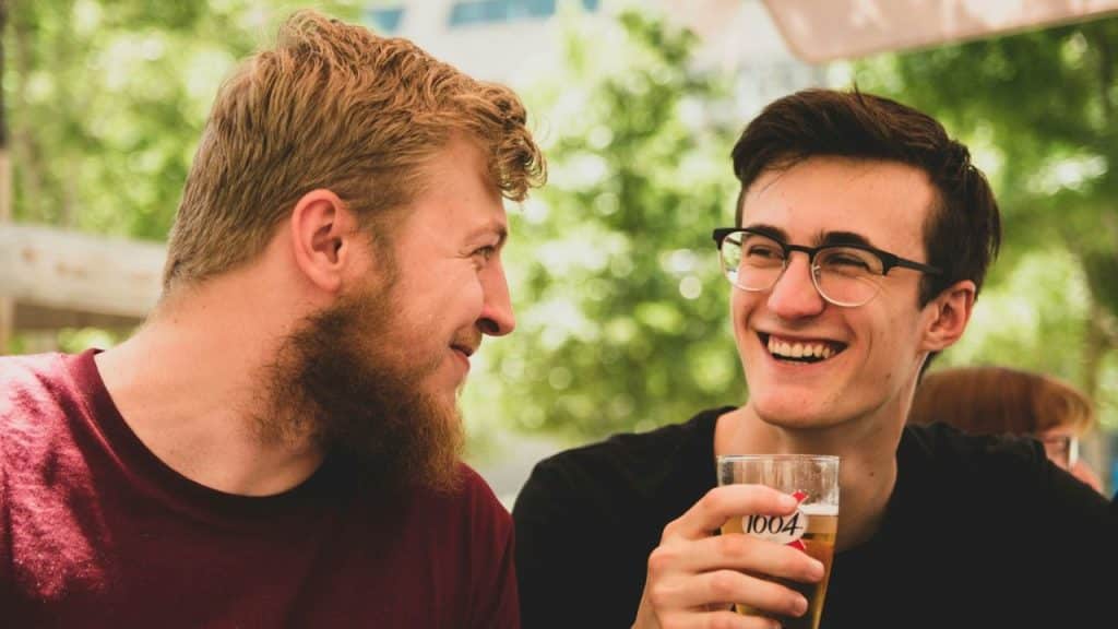 Two young men, one bearded and one in glasses, share a laugh outdoors.