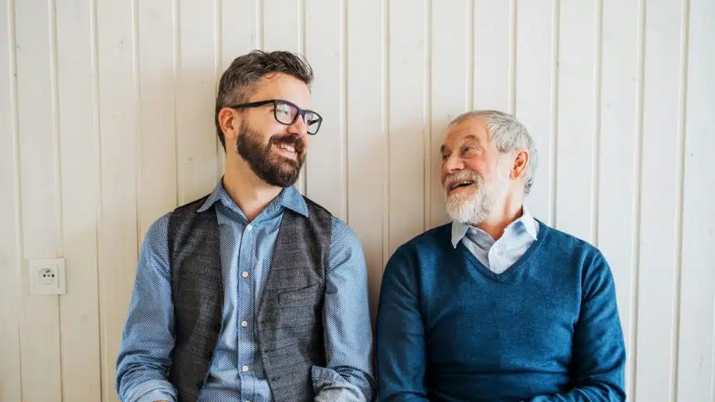 Two smiling men, one younger with glasses and one older with a beard, stand against a white wall.