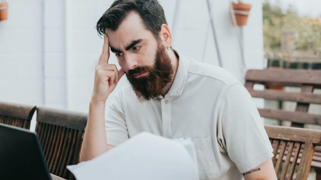 A bearded man sits outdoors, concentrating on papers with his hand pressed to his temple.