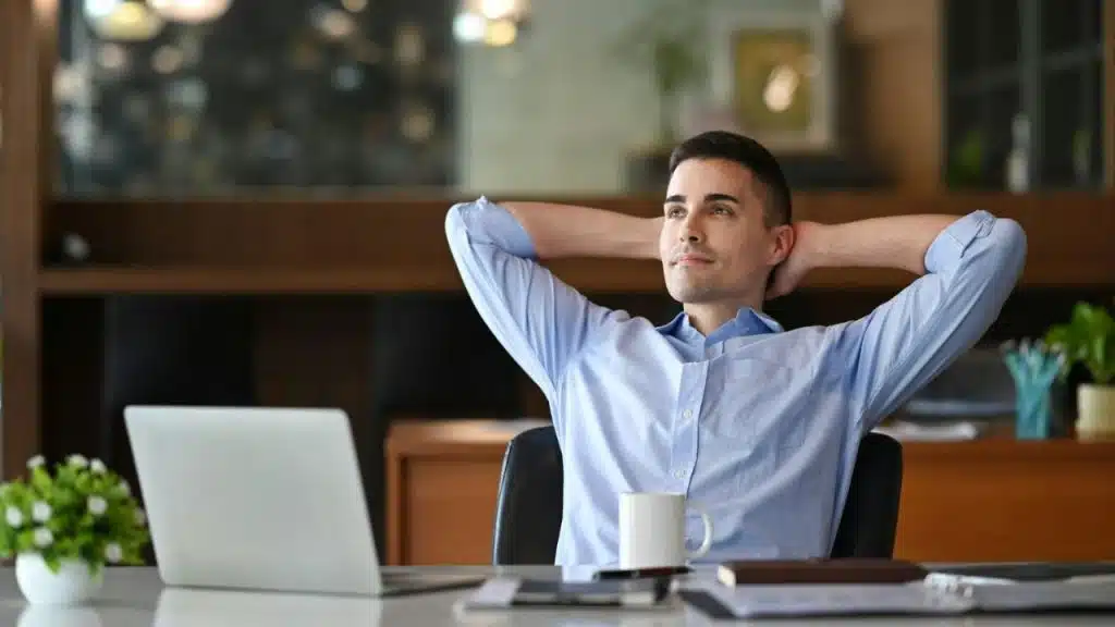 A man in a blue shirt relaxes with hands behind his head at a desk with a laptop.