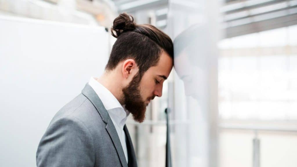 Man with a beard and a top knot, wearing a gray suit, leaning his forehead against a wall in a bright, modern space.