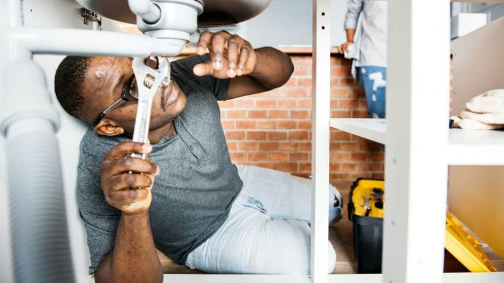 A man with glasses uses a wrench to work on plumbing under a sink.