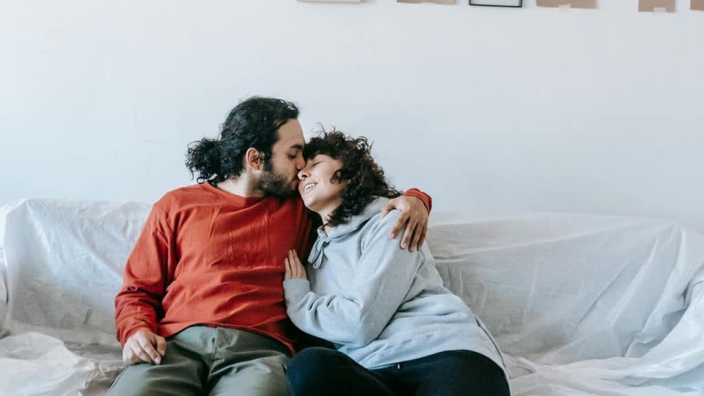A couple sitting on a couch while the man kisses the woman’s forehead.