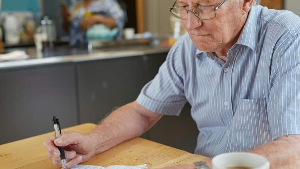 Older man in glasses and a striped shirt writing on paper at a table, with a coffee cup nearby.