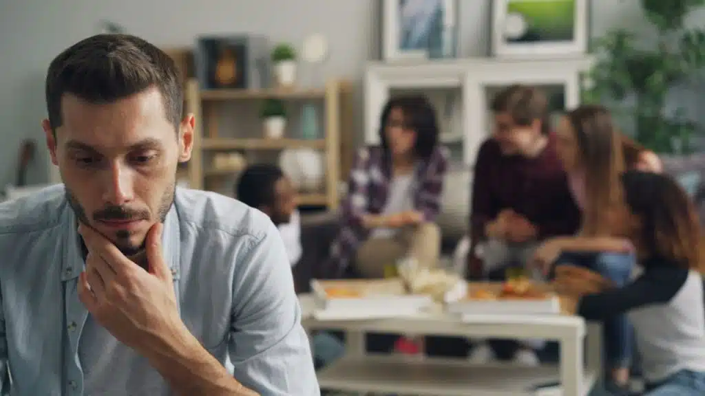 A pensive man is sitting in the foreground, while a group of friends talk in the background.
