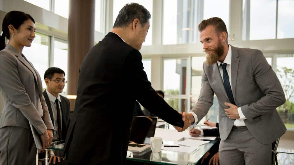 Two businessmen in suits shaking hands while other professionals observe in a modern office.