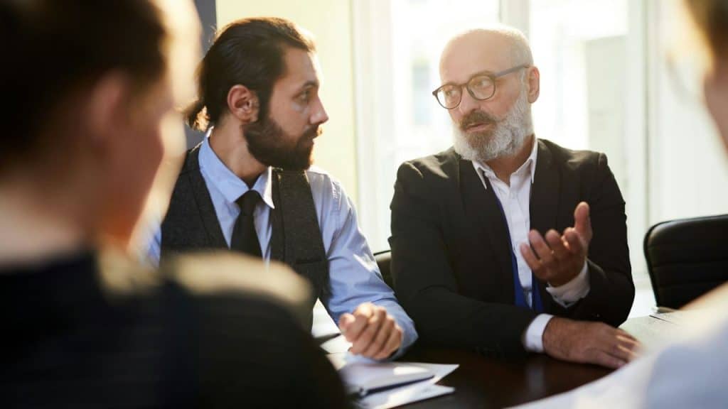 Two bearded men in professional attire discussing at a table in a bright room.