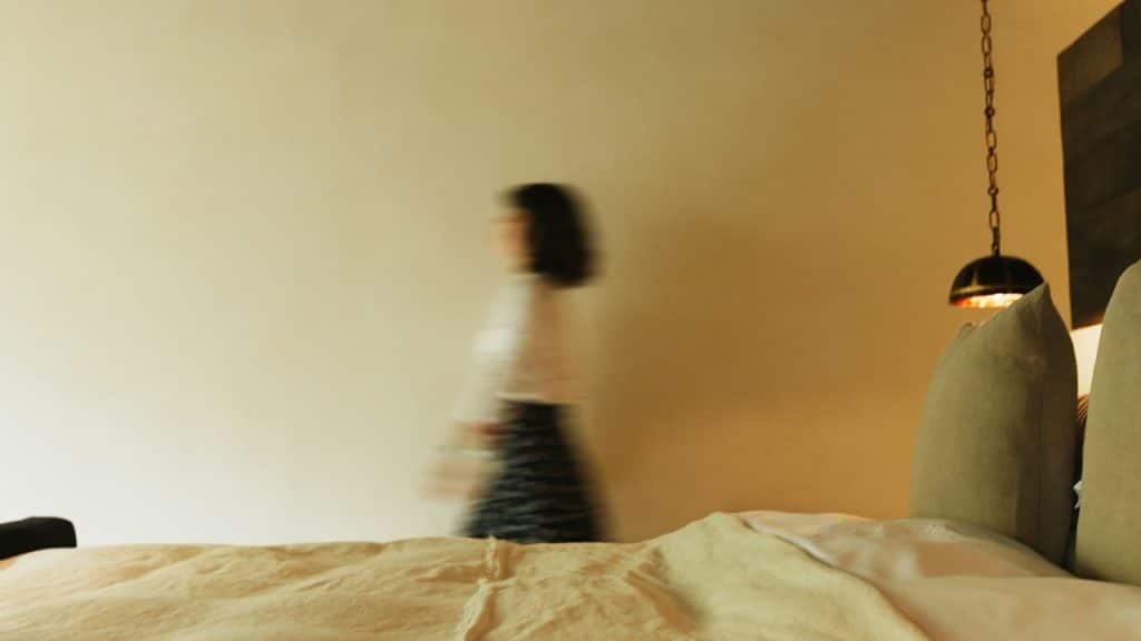 A blurry, long-exposure shot of a person walking past a bare wall behind the foot of a bed.