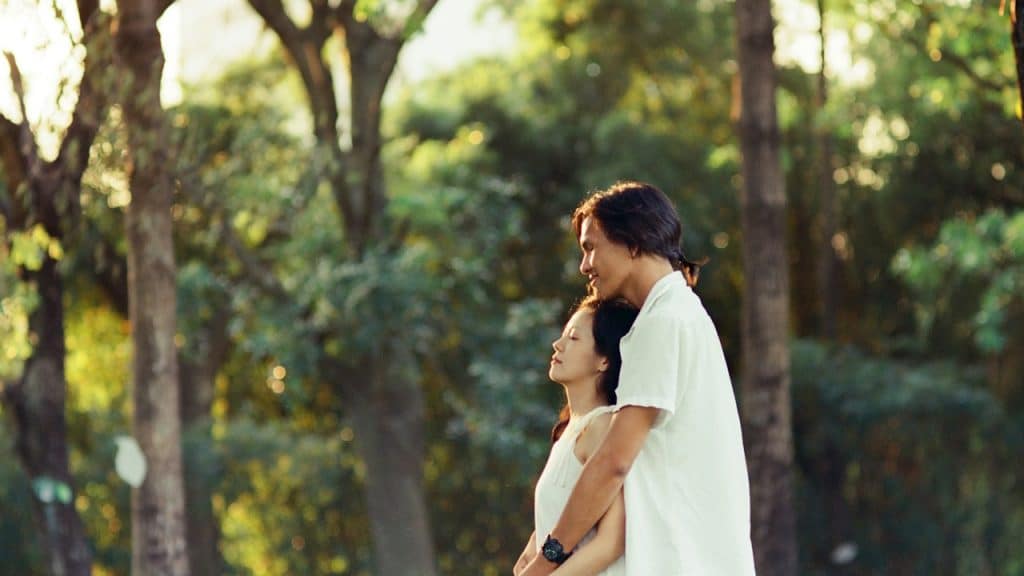 A tall man embraces a woman from behind in a sun-dappled park, with the woman closing her eyes peacefully.