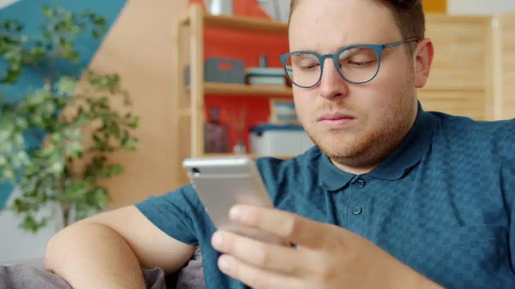 A man with red hair and blue glasses intently looks down at his smartphone.
