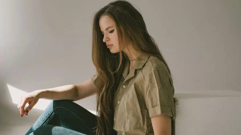 A young woman with long brown hair sits against a white wall, looking down with a sad expression.