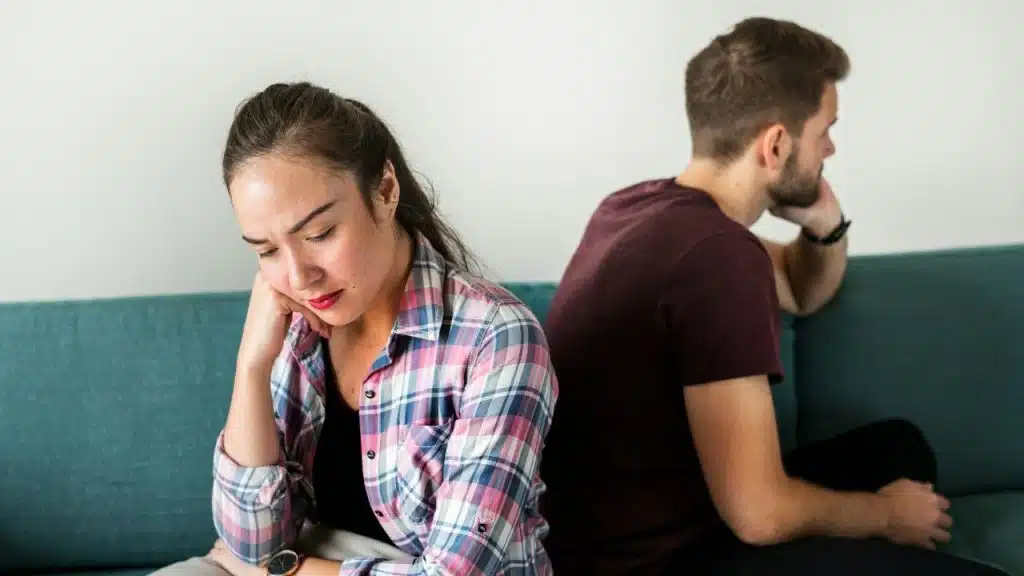 A man and woman sit on a teal couch, back-to-back, both looking distressed or withdrawn.
