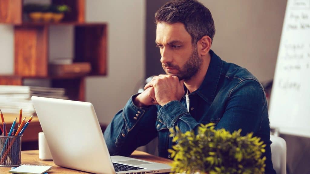 A serious man with his chin resting on his hands is looking intently at a laptop on a wooden desk.