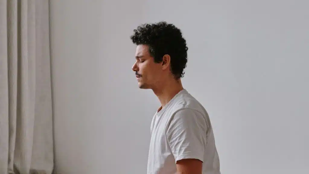 A man with curly hair and a mustache sits in profile against a plain white wall, eyes closed.
