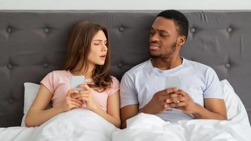 A Caucasian woman is staring suspiciously at an African-American man, both holding cell phones in bed.