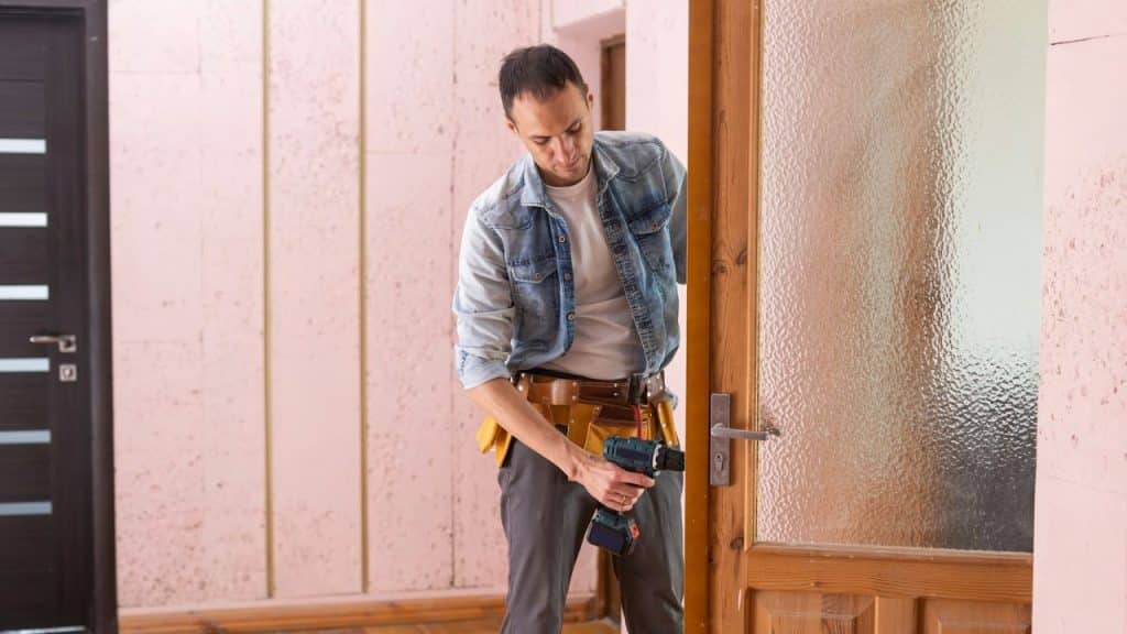 Man in a denim shirt uses a drill on a wooden door with a frosted glass panel.