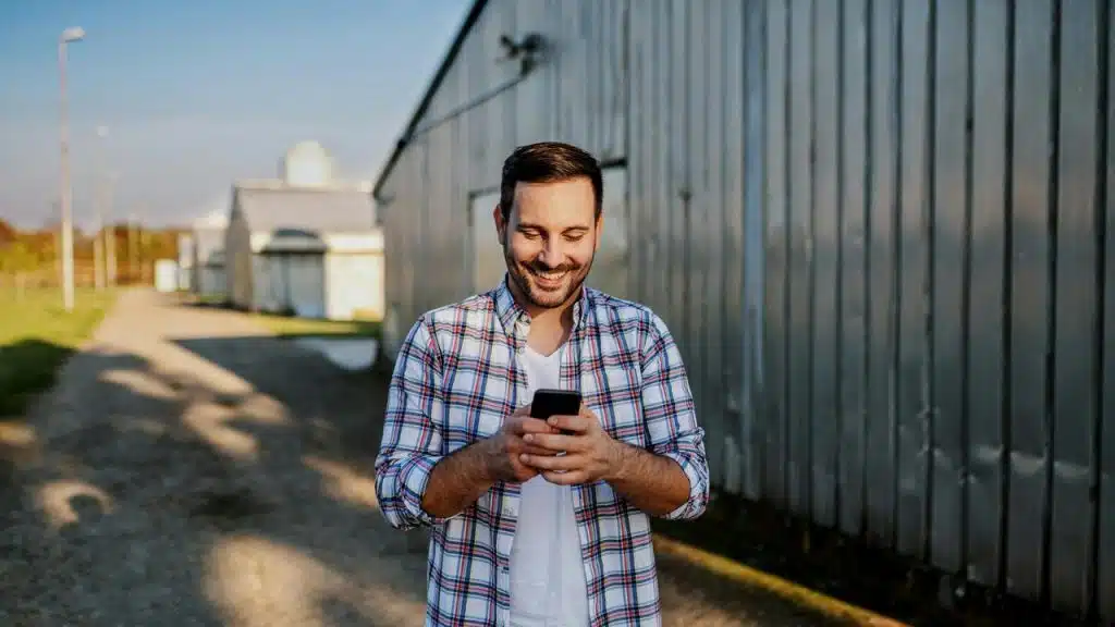 A man in a plaid shirt smiling while looking at his phone outdoors.