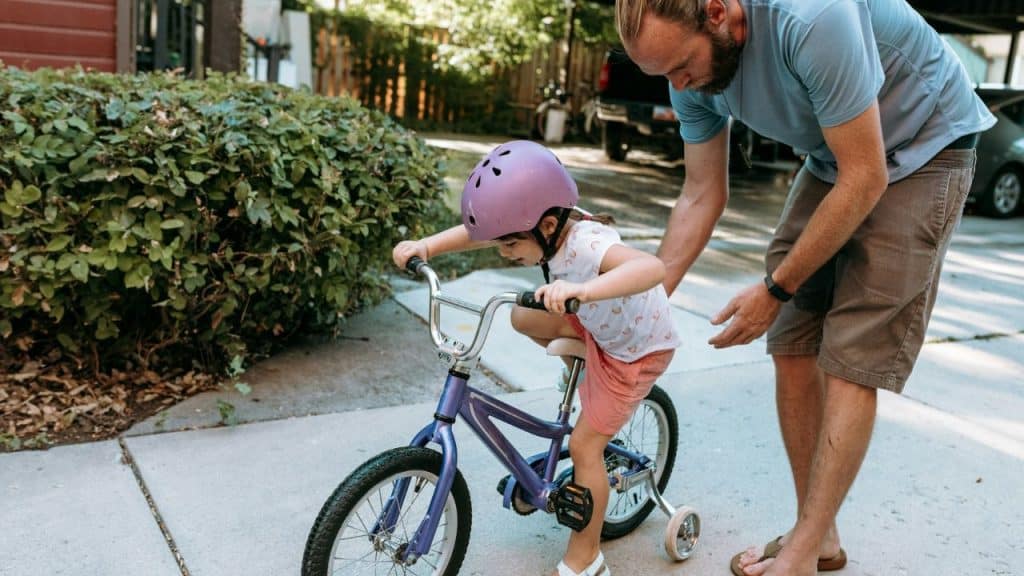 A man helps a young child in a purple helmet learn to ride a bicycle with training wheels on a sidewalk.