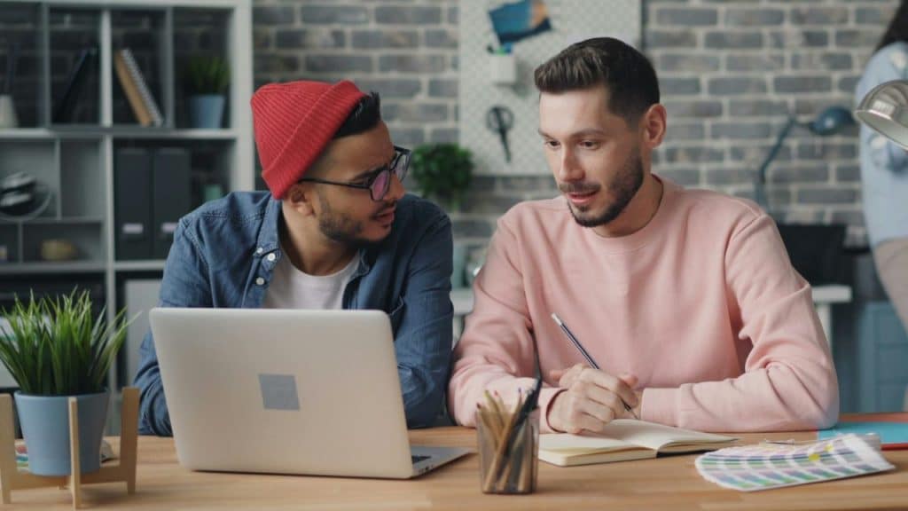 Two men at a desk look at a laptop, one holding a pencil and the other wearing a red beanie.