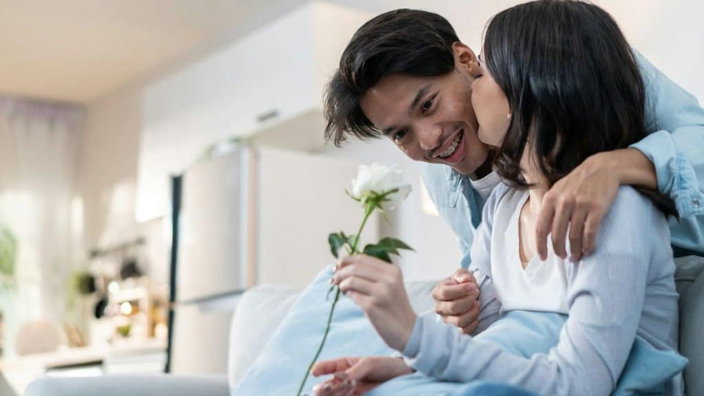 Young man putting his arm around a woman holding a white rose on a couch.