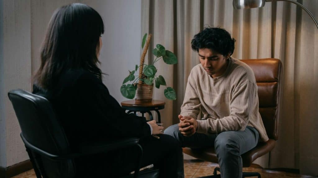 Young man looking down with clasped hands across from a woman in a black chair.