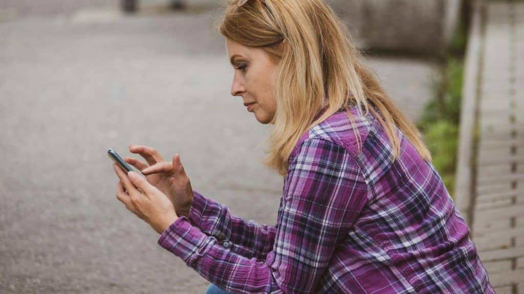 A woman sitting outdoors wearing a purple plaid shirt and using her smartphone.