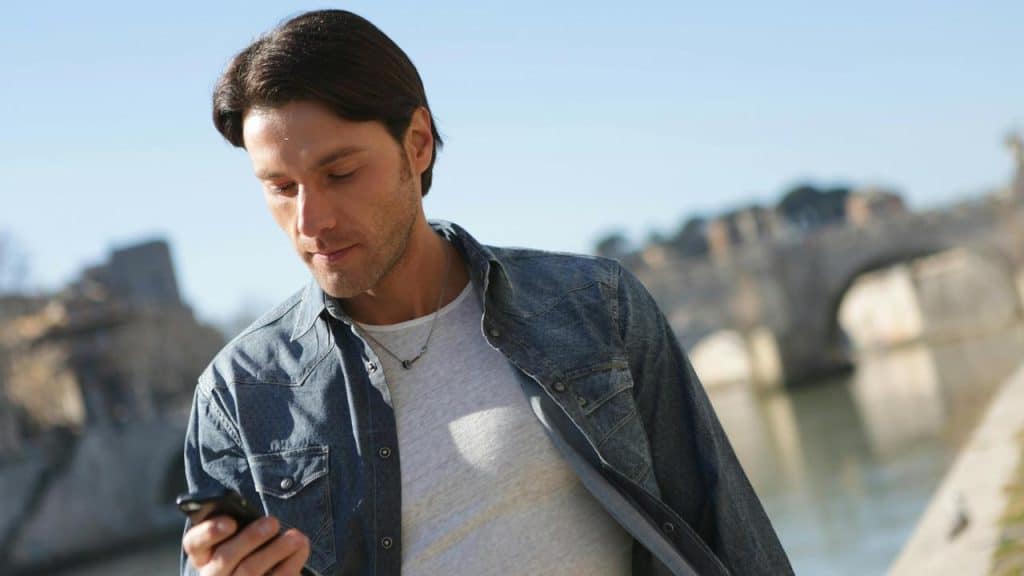A man outdoors looking at his phone near a river and an old stone bridge.