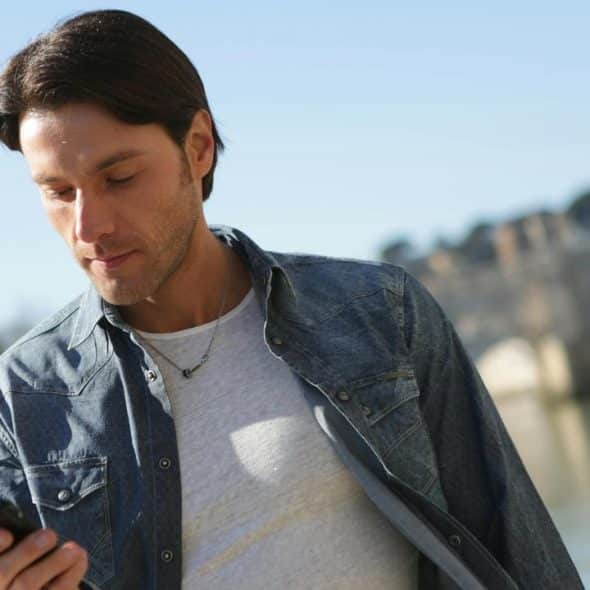 A man outdoors looking at his phone near a river and an old stone bridge.