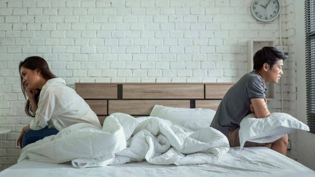 A man and woman sit separately on a bed with unmade white sheets, facing away from each other against a white brick wall.