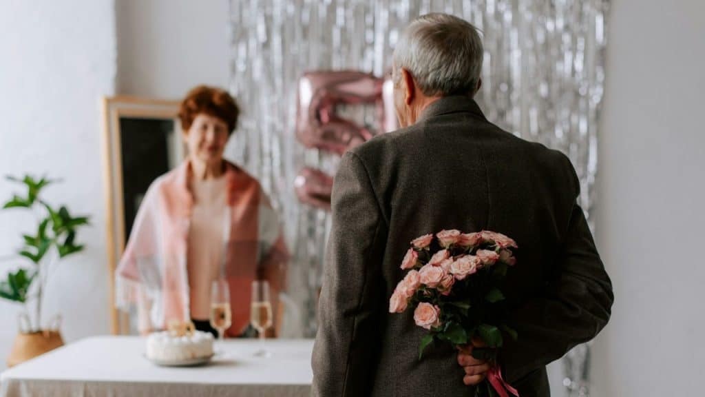 An older man stands with his back to the camera, holding a bouquet of pink roses behind his back for a woman smiling across a table.