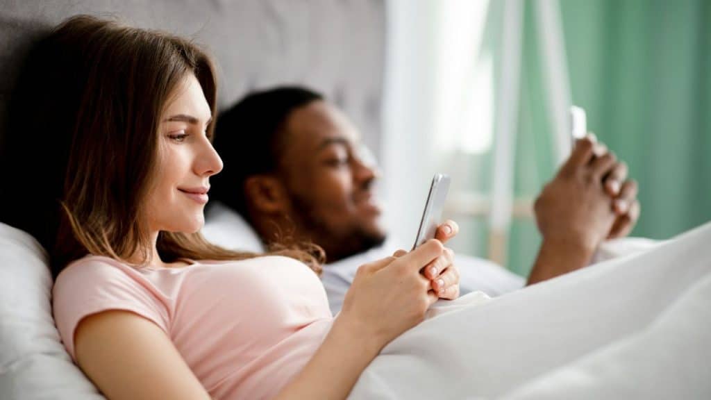 A couple lying in bed, each looking at their own smartphone.