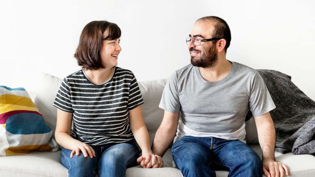 A casually dressed couple sits on a couch holding hands, smiling at each other.